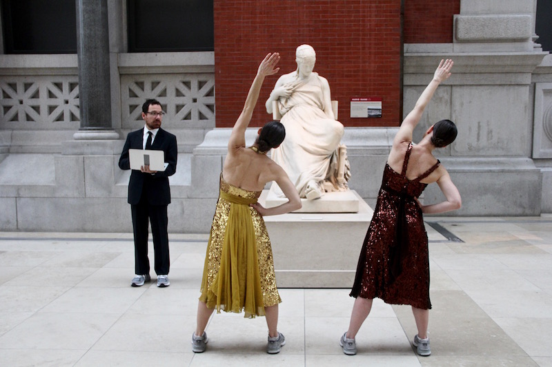 Two dancers stretch in front of a statue as a man in sneakers and a tux looks on with a laptop in his hand